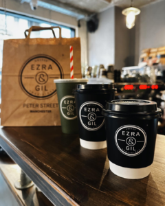 A photo of Ezra & Gil branded coffee cups with one of their brown paper branded bags in the background.