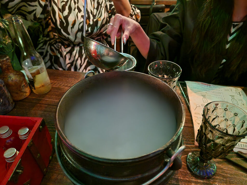 A steaming cauldron being stirred on a wooden table with drinking glasses surrounding it
