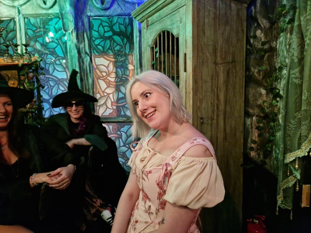A lady with blonde hair dressed in a pale pink dress and corset gives an eerie smile, while two witches in black cloaks and hats laugh in the background