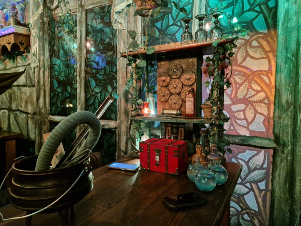 A wooden table against a stained glass effect background, with a red box in front of a display of curious items. A cauldron with ladle sits in the foreground.