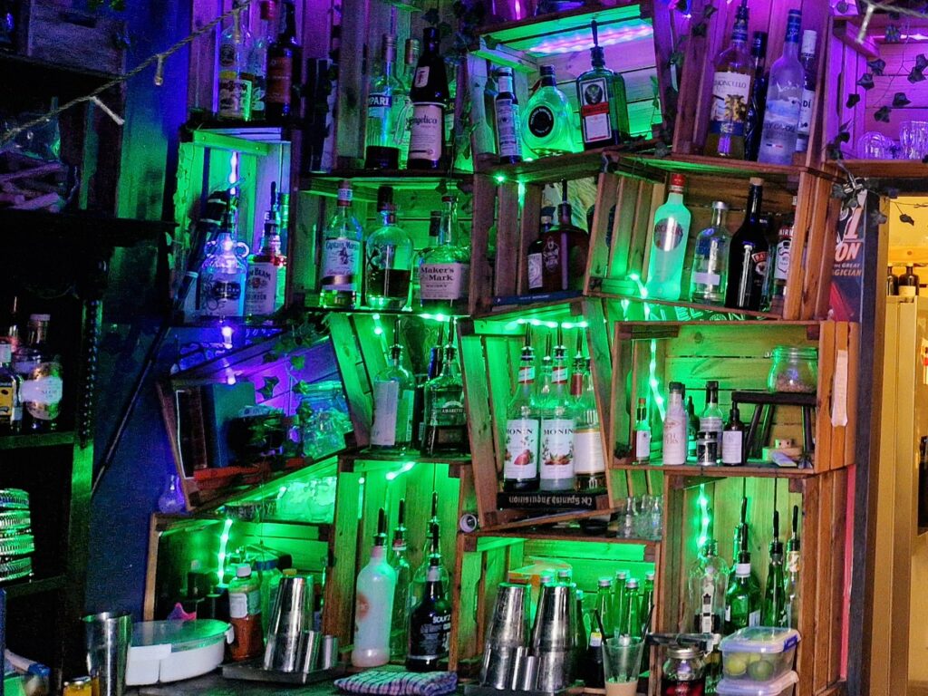 Bottles of spirits and syrups illuminated with neon green lighting, stacked in wonky wooden crates