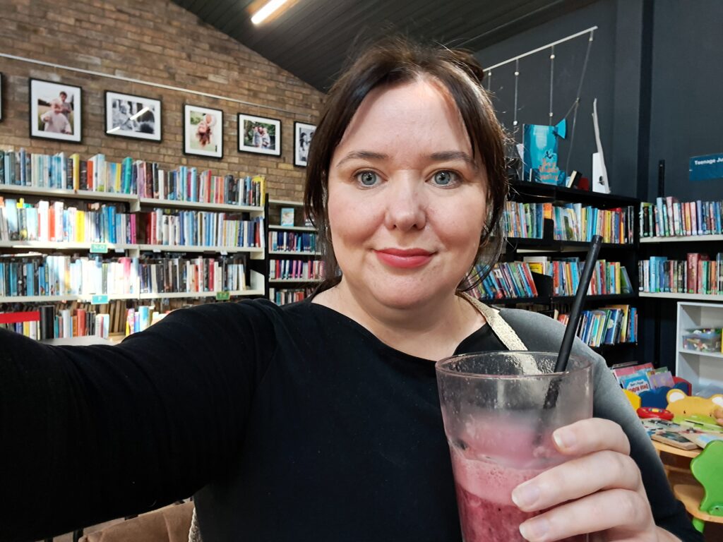 Photograph of this article's author holding a blueberry smoothie against the backdrop of the library space.