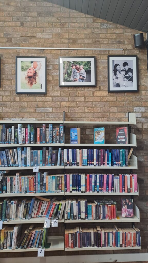 Photograph of library bookcases against a light brick wall. Three framed photographs are placed above the books.