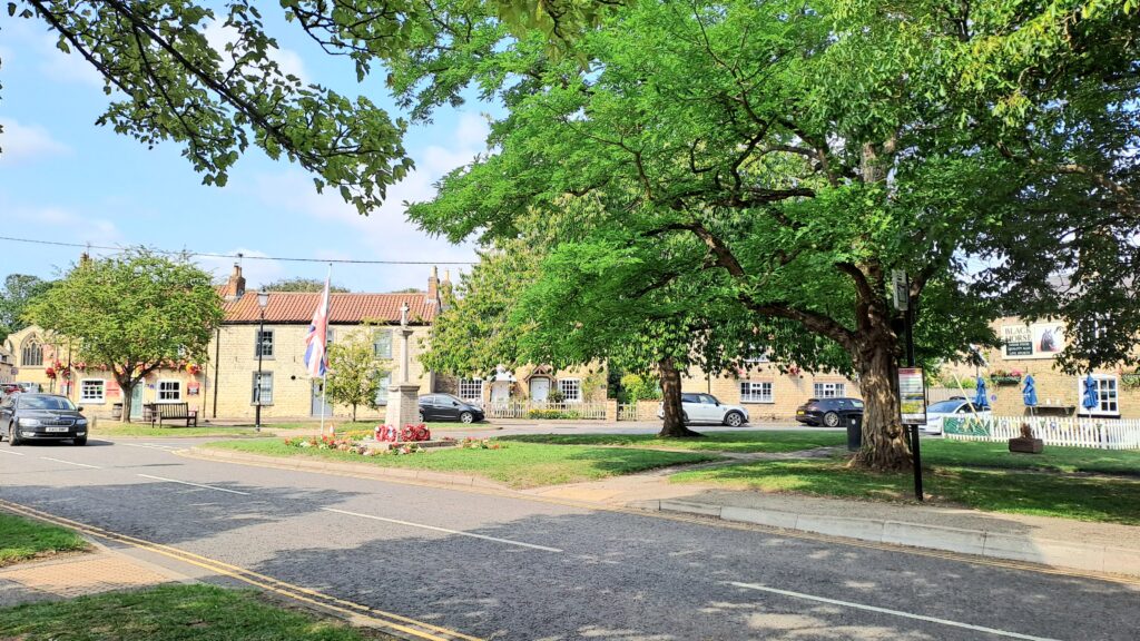 Photograph of Nettleham village green, featuring two large green trees, stone coloured buildings and a bus stop.