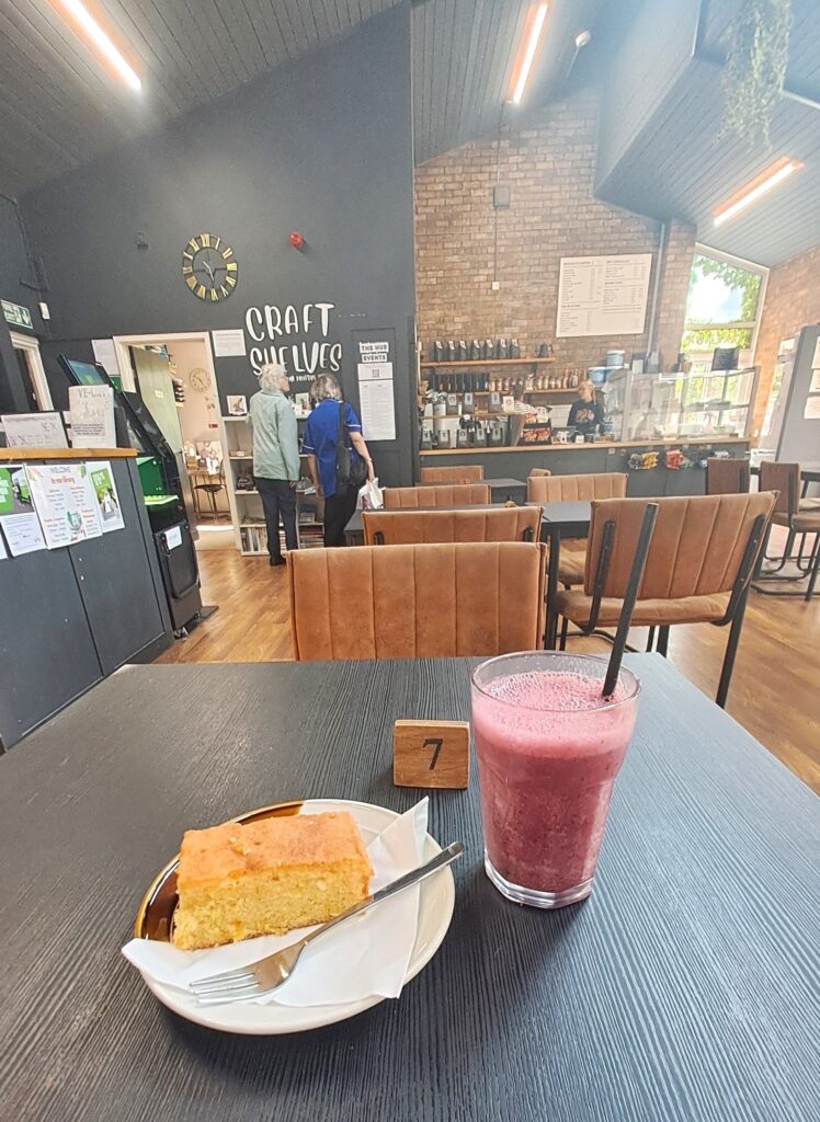 Photograph of Nettleham Community Hub's library-café space. In the foreground is a slice of lemon drizzle cake and a purple smoothie on a black table.